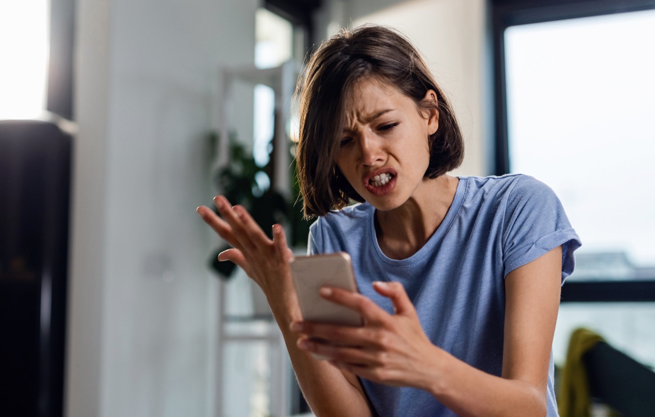 Young frustrated woman reading text message cell phone in disbelief while sitting on the bed at home.
